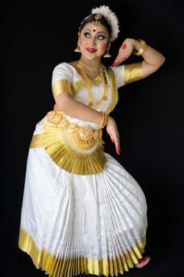 A woman is dressed in traditional Indian dance attire, featuring a white and gold pleated skirt, ornate jewelry, and a decorative headpiece. Her face is adorned with makeup, and her hands show expressive gestures with red markings on her palms. She is posed against a dark background which emphasizes her colorful costume.