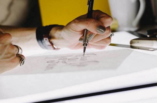 A hand is holding a mechanical pencil, sketching intricate designs on a sheet of paper. The image is brightly lit, with focus on the detailed work and the artistic process. Surrounding the hand are other drawing tools like a ruler and a utility knife.