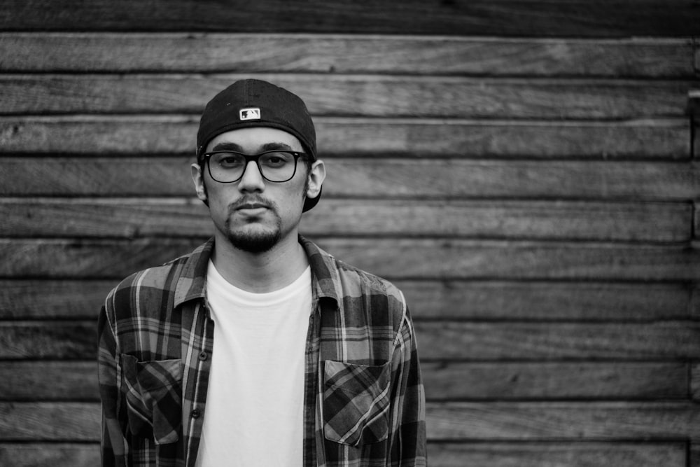 grayscale photo of man in plaid shirt and cap standing in front of wooden panel