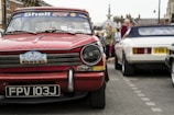 A classic red Triumph car with a license plate reading FPV 103J is prominently displayed. The windshield is decorated with Shell Sport and other stickers. In the background, people are gathering around other vintage cars, including a white one. The street setting shows signs of a car event or exhibition.