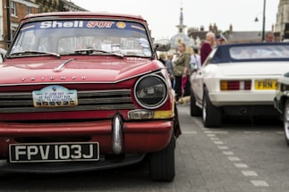 A classic red Triumph car with a license plate reading FPV 103J is prominently displayed. The windshield is decorated with Shell Sport and other stickers. In the background, people are gathering around other vintage cars, including a white one. The street setting shows signs of a car event or exhibition.