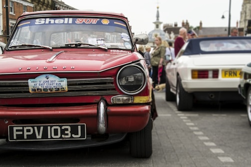 A classic red Triumph car with a license plate reading FPV 103J is prominently displayed. The windshield is decorated with Shell Sport and other stickers. In the background, people are gathering around other vintage cars, including a white one. The street setting shows signs of a car event or exhibition.