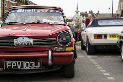 A classic red Triumph car with a license plate reading FPV 103J is prominently displayed. The windshield is decorated with Shell Sport and other stickers. In the background, people are gathering around other vintage cars, including a white one. The street setting shows signs of a car event or exhibition.