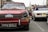A classic red Triumph car with a license plate reading FPV 103J is prominently displayed. The windshield is decorated with Shell Sport and other stickers. In the background, people are gathering around other vintage cars, including a white one. The street setting shows signs of a car event or exhibition.