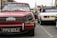 A classic red Triumph car with a license plate reading FPV 103J is prominently displayed. The windshield is decorated with Shell Sport and other stickers. In the background, people are gathering around other vintage cars, including a white one. The street setting shows signs of a car event or exhibition.