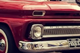A close-up view of the front of a vintage red Chevrolet vehicle, featuring a classic design with prominent headlights, chrome detailing, and a visible license plate with the number FKKI03C. The paint is glossy, reflecting the surroundings, and the car's brand name is displayed on the grille.