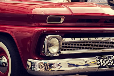 A close-up view of the front of a vintage red Chevrolet vehicle, featuring a classic design with prominent headlights, chrome detailing, and a visible license plate with the number FKKI03C. The paint is glossy, reflecting the surroundings, and the car's brand name is displayed on the grille.