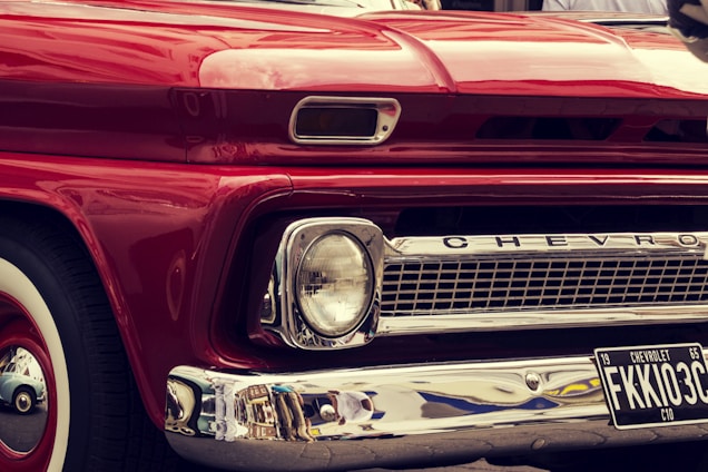A close-up view of the front of a vintage red Chevrolet vehicle, featuring a classic design with prominent headlights, chrome detailing, and a visible license plate with the number FKKI03C. The paint is glossy, reflecting the surroundings, and the car's brand name is displayed on the grille.