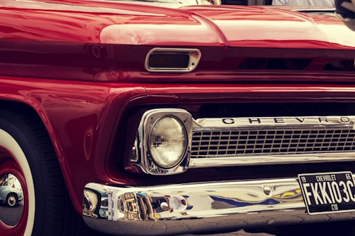 A close-up view of the front of a vintage red Chevrolet vehicle, featuring a classic design with prominent headlights, chrome detailing, and a visible license plate with the number FKKI03C. The paint is glossy, reflecting the surroundings, and the car's brand name is displayed on the grille.