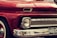 A close-up view of the front of a vintage red Chevrolet vehicle, featuring a classic design with prominent headlights, chrome detailing, and a visible license plate with the number FKKI03C. The paint is glossy, reflecting the surroundings, and the car's brand name is displayed on the grille.
