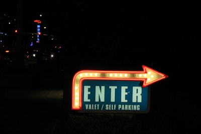 A brightly lit sign with an arrow pointing to the right, indicating 'ENTER' for valet or self parking. The sign is illuminated with white and red lights and is placed in a dark environment with city lights in the background.