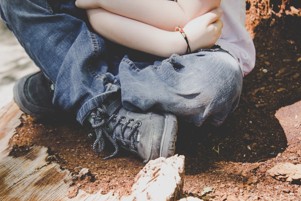 A child in jeans, boots, and a bracelet sits on dirt, crossing their legs