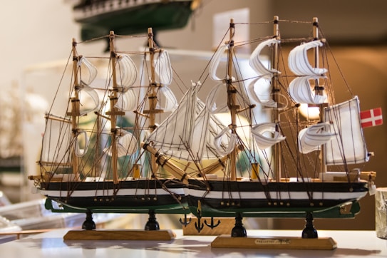 A detailed model ship with multiple masts and sails, intricately crafted with strings representing rigging. The model is displayed indoors with warm lighting accentuating its elegant details, and a small flag is attached at the rear.