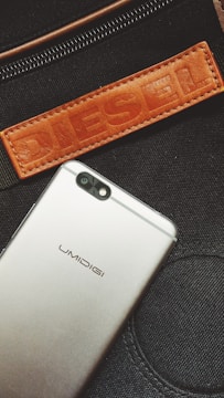 A gray smartphone with the UMIDIGI logo is positioned diagonally over a black fabric background. Above the phone, a brown leather patch with the word 'DIESEL' embossed on it is seen, along with a zipper detail.