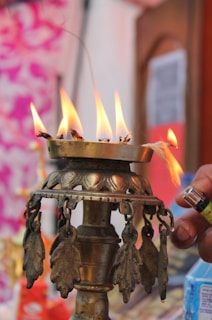 A traditional metal lamp with multiple flames is burning, likely used for religious or ceremonial purposes. The lamp has intricate designs and metal decorations hanging from it. A hand holding a lighter is visible, suggesting the act of lighting the lamp. The background is blurred, with hints of colorful patterns.
