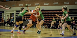 A group of women are playing an intense game of handball indoors. One player in a red and white jersey is in possession of the ball, guarded closely by two opposing players in green jerseys. Spectators and team members are seated in the background on benches. The floor is a polished wooden court with blue and white markings.