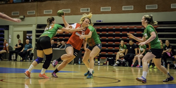 A group of women are playing an intense game of handball indoors. One player in a red and white jersey is in possession of the ball, guarded closely by two opposing players in green jerseys. Spectators and team members are seated in the background on benches. The floor is a polished wooden court with blue and white markings.