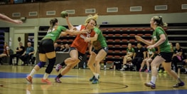 A group of women are playing an intense game of handball indoors. One player in a red and white jersey is in possession of the ball, guarded closely by two opposing players in green jerseys. Spectators and team members are seated in the background on benches. The floor is a polished wooden court with blue and white markings.
