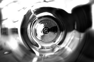 A black and white close-up image looking through the bottom of a glass bottle. The circular base at the center has an embossed letter 'H' surrounded by intricate patterns. Light and shadows create a dramatic effect within the curved and reflective surfaces.