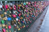 A dense collection of colorful padlocks attached to a wire fence on a bridge, commonly known as love locks. The locks are various sizes and colors, including red, blue, and yellow. They symbolize promises of love and commitment.