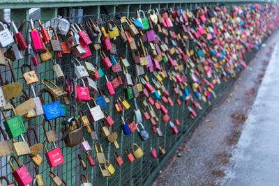 A dense collection of colorful padlocks attached to a wire fence on a bridge, commonly known as love locks. The locks are various sizes and colors, including red, blue, and yellow. They symbolize promises of love and commitment.