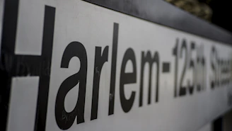 A close-up view of a sign with the text 'Harlem-125th Street', featuring large black letters on a white background, slightly out of focus towards the end.