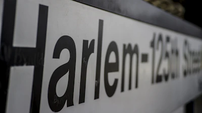 A close-up view of a sign with the text 'Harlem-125th Street', featuring large black letters on a white background, slightly out of focus towards the end.
