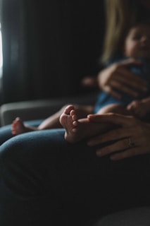 A close-up view of a baby's feet resting on an adult's lap, with the adult's hands gently holding or caressing the child. The setting appears to be dimly lit, creating a soft, intimate ambiance.