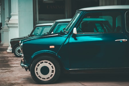 A row of vintage cars, primarily a dark teal Mini Cooper, is lined up along a street with classical architecture in the background. The cars have a shiny finish, highlighting the retro style, and are parked in front of a building that has white columns.