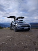 A black Tesla SUV with its falcon-wing doors open is parked on a dirt path. Behind the car, there is a scenic view of mountains and a cloudy sky.