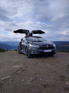 A black Tesla SUV with its falcon-wing doors open is parked on a dirt path. Behind the car, there is a scenic view of mountains and a cloudy sky.