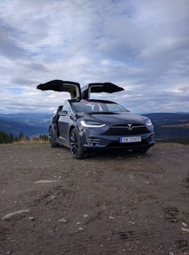 A black Tesla SUV with its falcon-wing doors open is parked on a dirt path. Behind the car, there is a scenic view of mountains and a cloudy sky.