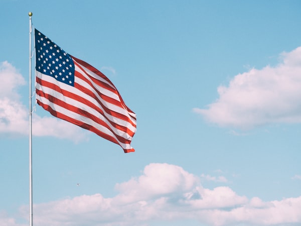 Flag of the United States of America on a flagpole, in front of a blue sky with white clouds.