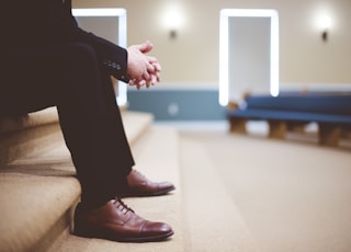 man in black pants and pair of brown leather lace-up shoes sitting on brown carpeted stairs inside room