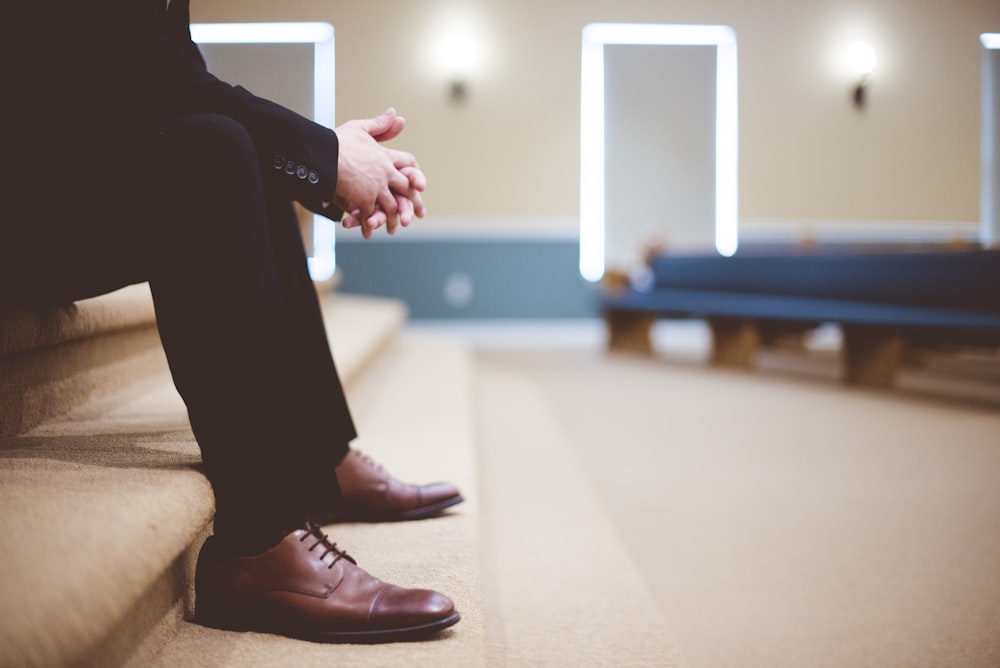 man in black pants and pair of brown leather lace-up shoes sitting on brown carpeted stairs inside room