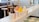 Four cups of soft-serve ice cream in various colors, including chocolate, pink, yellow, and white, are lined up on a counter. Each cup is labeled as 'Dairy, Fat, and Gluten Free.' The background features a cozy, retro-style interior with stools and checkered floor tiles.