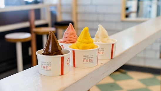 Four cups of soft-serve ice cream in various colors, including chocolate, pink, yellow, and white, are lined up on a counter. Each cup is labeled as 'Dairy, Fat, and Gluten Free.' The background features a cozy, retro-style interior with stools and checkered floor tiles.