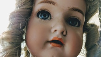 A close-up view of a vintage porcelain doll face with large, glassy blue eyes and finely painted eyebrows. The doll’s lips are painted a vibrant red, and it has curly blonde hair. The focus is on the intricate details of the doll’s facial features.