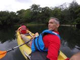 A man in a red and blue life jacket is kayaking on a calm body of water surrounded by lush green forest. Another person wearing a red shirt and life jacket sits in front of him in the kayak. The environment appears serene and natural.