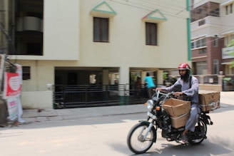A man wearing a red helmet is riding a motorcycle on a city street. The motorcycle is loaded with large cardboard boxes. The background shows a building with several windows and a driveway, and another person is visible walking in the background.