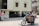 A man wearing a red helmet is riding a motorcycle on a city street. The motorcycle is loaded with large cardboard boxes. The background shows a building with several windows and a driveway, and another person is visible walking in the background.