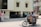A man wearing a red helmet is riding a motorcycle on a city street. The motorcycle is loaded with large cardboard boxes. The background shows a building with several windows and a driveway, and another person is visible walking in the background.