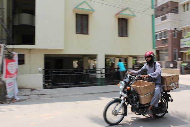 A man wearing a red helmet is riding a motorcycle on a city street. The motorcycle is loaded with large cardboard boxes. The background shows a building with several windows and a driveway, and another person is visible walking in the background.