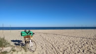 A bicycle loaded with camping gear rests on a sandy beach, facing the calm ocean under a clear blue sky. Several poles are visible in the distance, possibly marking boundaries or pathways.