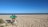 A bicycle loaded with camping gear rests on a sandy beach, facing the calm ocean under a clear blue sky. Several poles are visible in the distance, possibly marking boundaries or pathways.