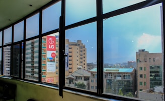A view through large glass windows showcasing a cityscape with several multi-story buildings. One of the buildings prominently displays an LG advertisement. The sky is clear with a hint of clouds, adding to the urban environment captured from a higher floor with a wide-angle perspective.