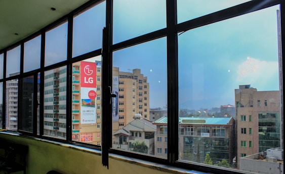 A view through large glass windows showcasing a cityscape with several multi-story buildings. One of the buildings prominently displays an LG advertisement. The sky is clear with a hint of clouds, adding to the urban environment captured from a higher floor with a wide-angle perspective.