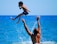 A child wearing blue swim shorts is playfully tossed into the air by an adult in the water against a bright blue ocean backdrop. The child's legs are bent, and their arms are close to their body, with a dynamic splash of water below.
