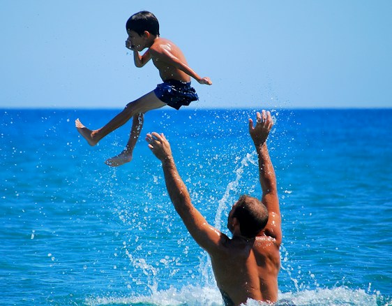 A child wearing blue swim shorts is playfully tossed into the air by an adult in the water against a bright blue ocean backdrop. The child's legs are bent, and their arms are close to their body, with a dynamic splash of water below.