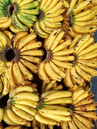 A large number of bunches of bananas are closely packed together, displaying varying shades of yellow and some green. The bananas have some brown specks and marks, indicating ripeness.